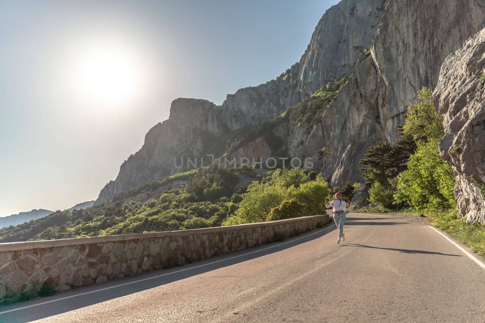 A woman runs along an asphalt road in the middle of a mountainous area. She is dressed in jeans and a white shirt, her hair is braided. A traveler in the mountains rejoices in nature