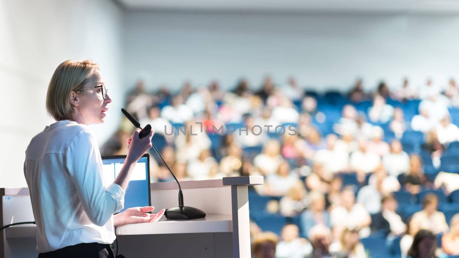 Female speaker giving a talk on corporate business conference. Unrecognizable people in audience at conference hall. Business and Entrepreneurship event