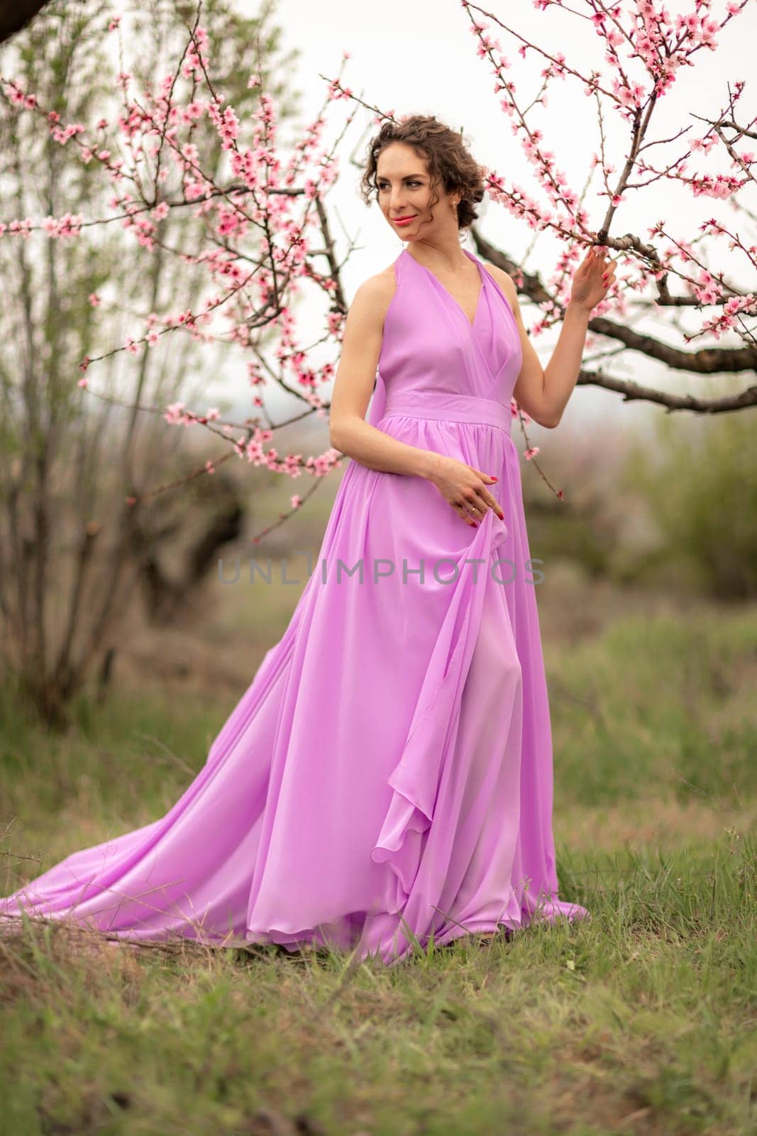 Woman peach blossom. Happy curly woman in pink dress walking in the garden of blossoming peach trees in spring by Matiunina