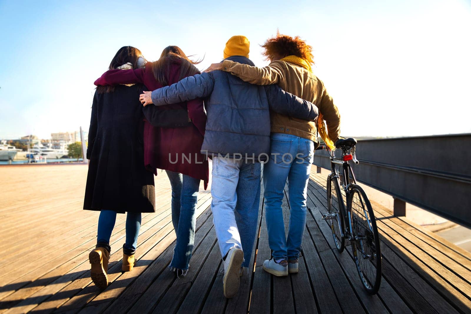 Rear view of group of friends walking together enjoying sunny winter afternoon.Multiracial friends embracing in the city by Hoverstock