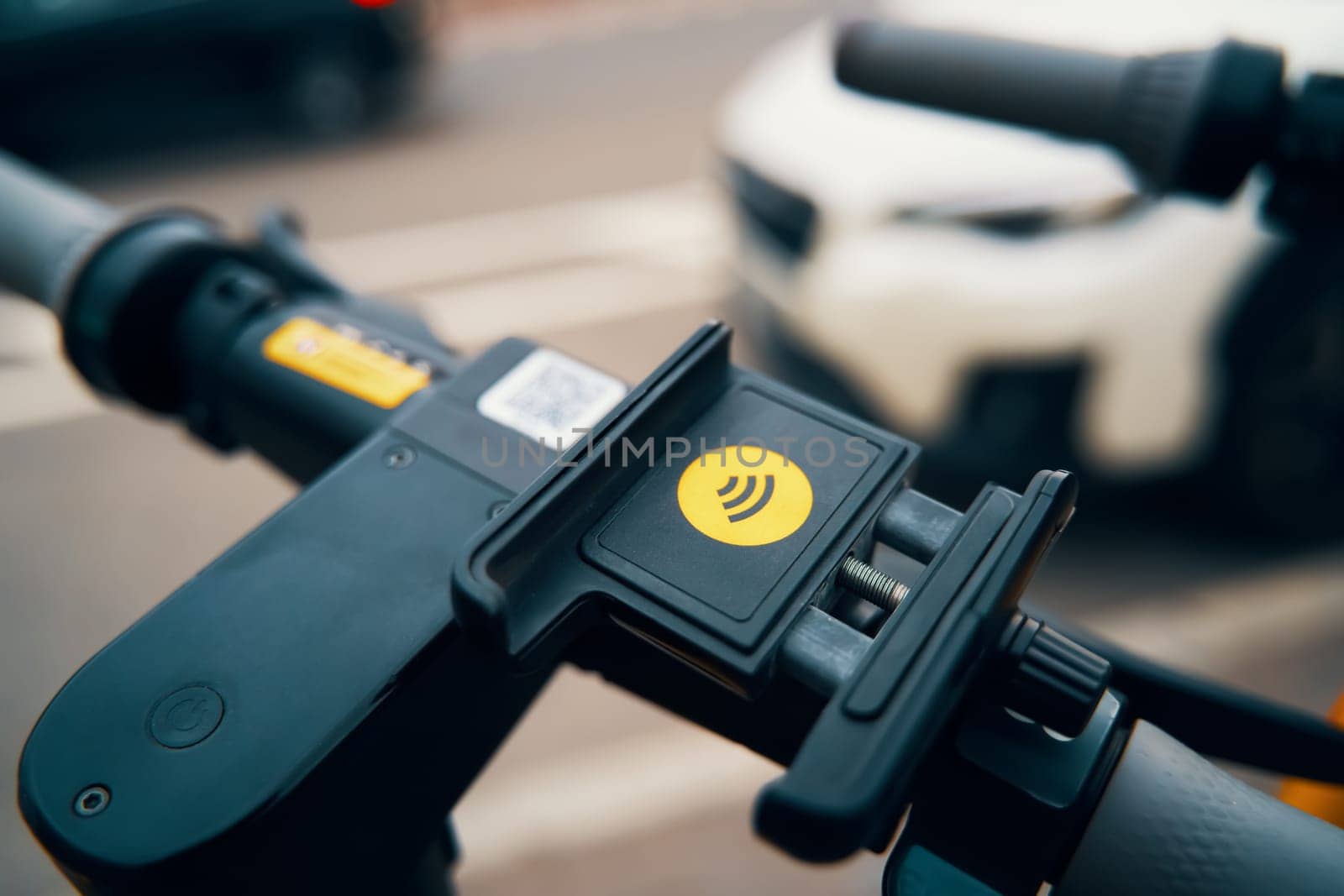 A close up of a phone holder on a scooter with a car in the background, resembling a small machine gun accessory on a larger automotive tire