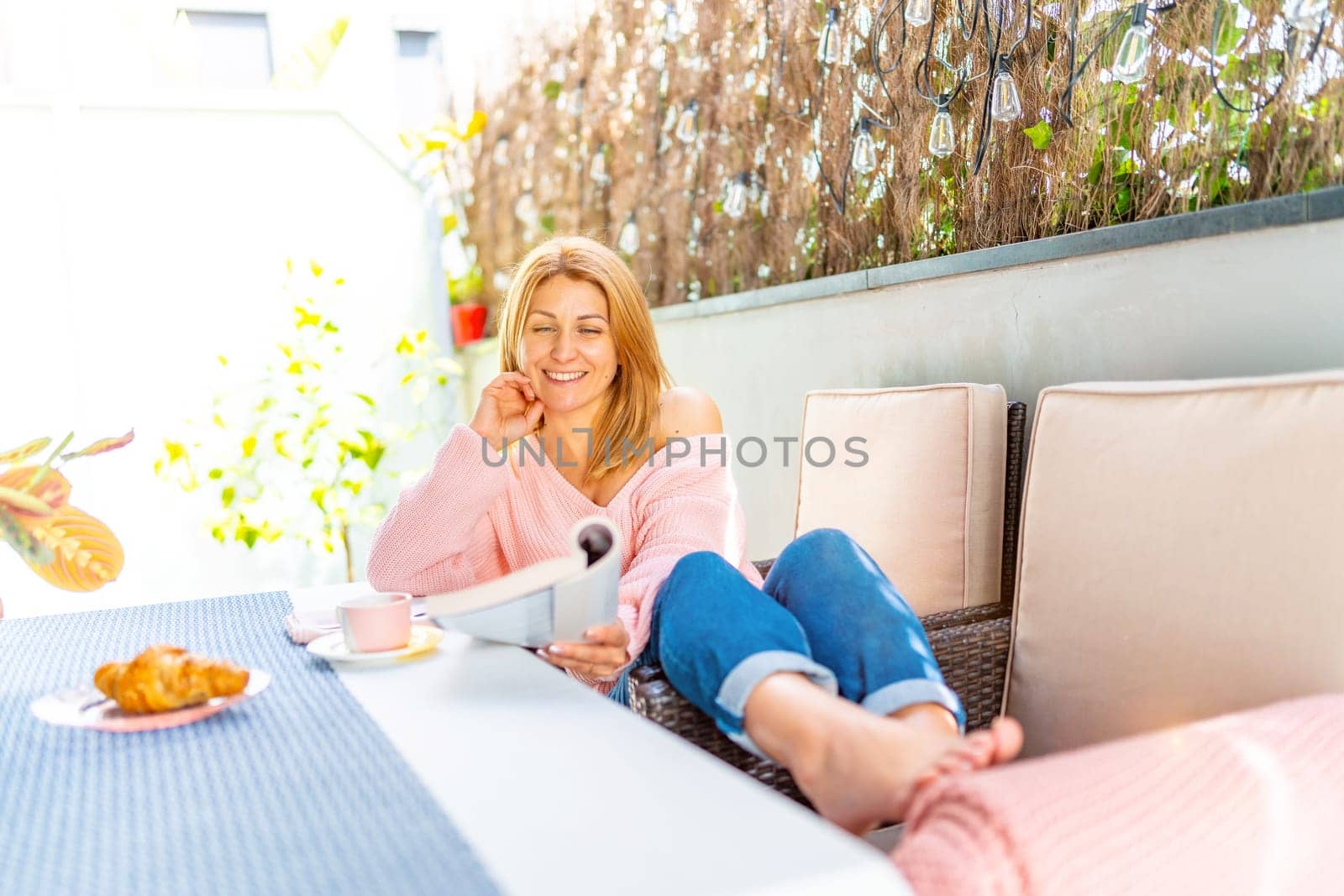 Relaxed caucasian adult blonde woman having breakfast while reading a book at the patio at home