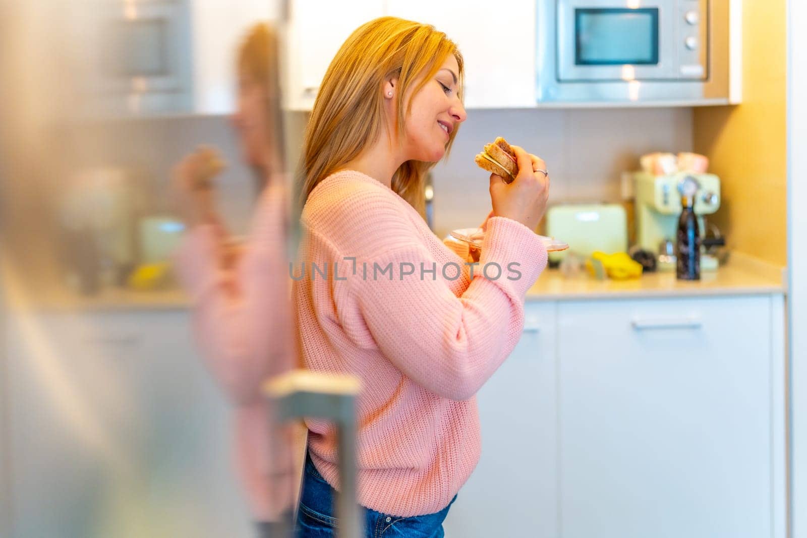 Side view of a caucasian blonde woman enjoying a delicious healthy sandwich standing in the kitchen at home