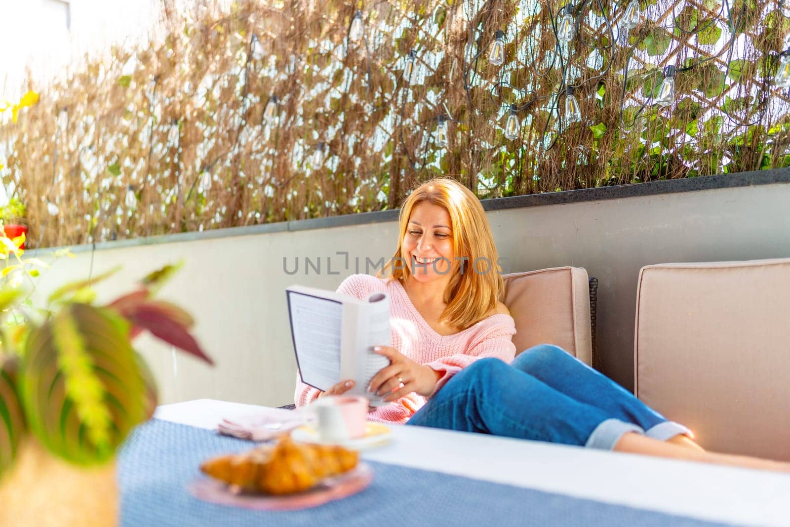 Caucasian adult blonde woman reading a book lying on outdoor sofa in the patio
