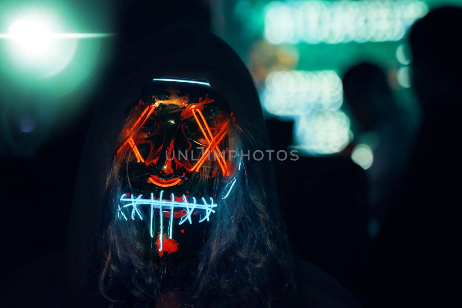 Human in hood with long disheveled hair in a lighting neon glowing mask. Anonymous man is a cyber criminal. Halloween dress-up party in night club. All Saints' Night.