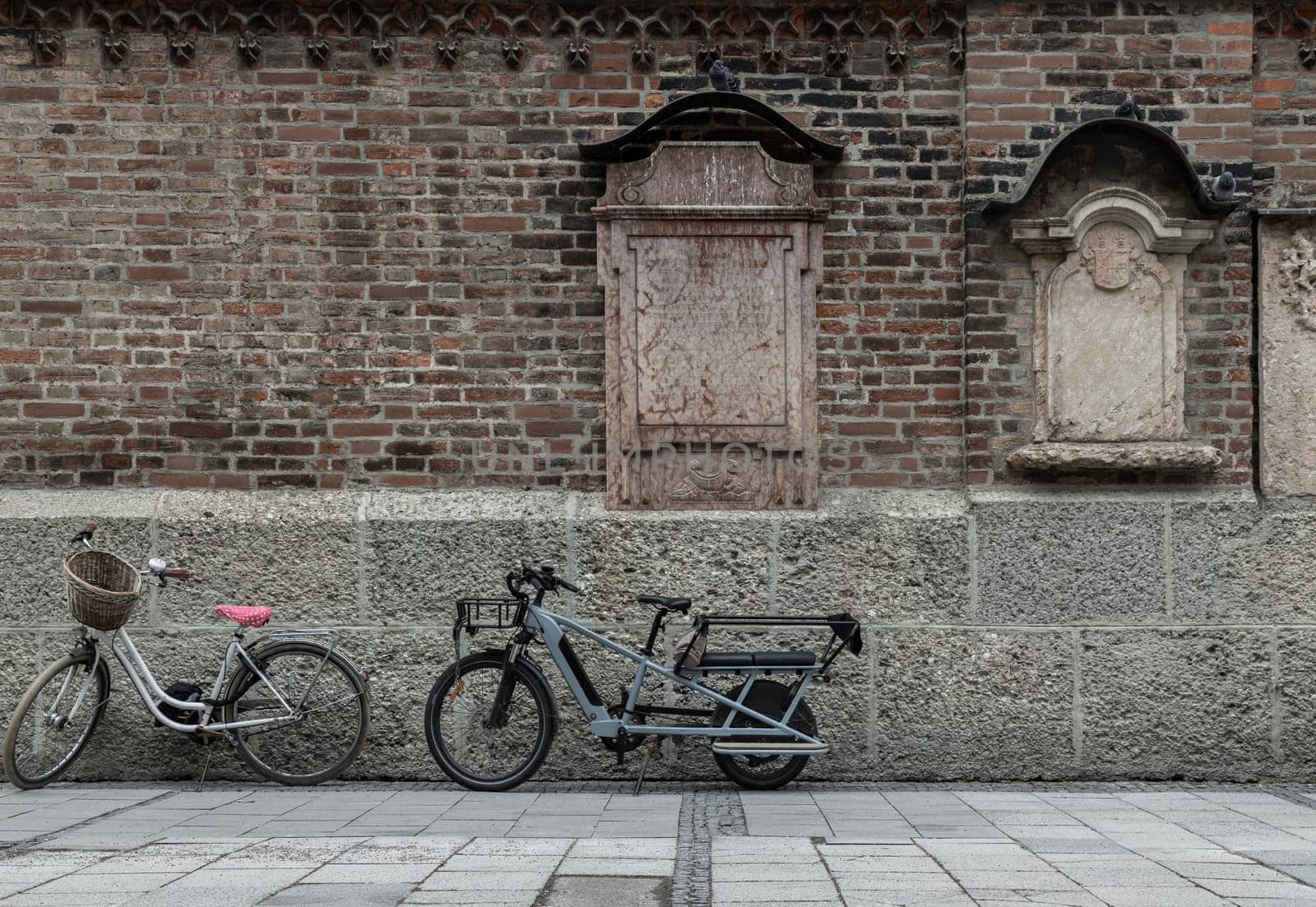Munich, Germany - Dec 21, 2023 - Vintage bike with basket and Electric longtail cargo bike are parked side of Mounted burial headstones based upon the outside Old brick wall of the Munich Frauenkirche or Frauenplatz (Cathedral Church of Our Lady) in Munich. Space for text, Selective focus.