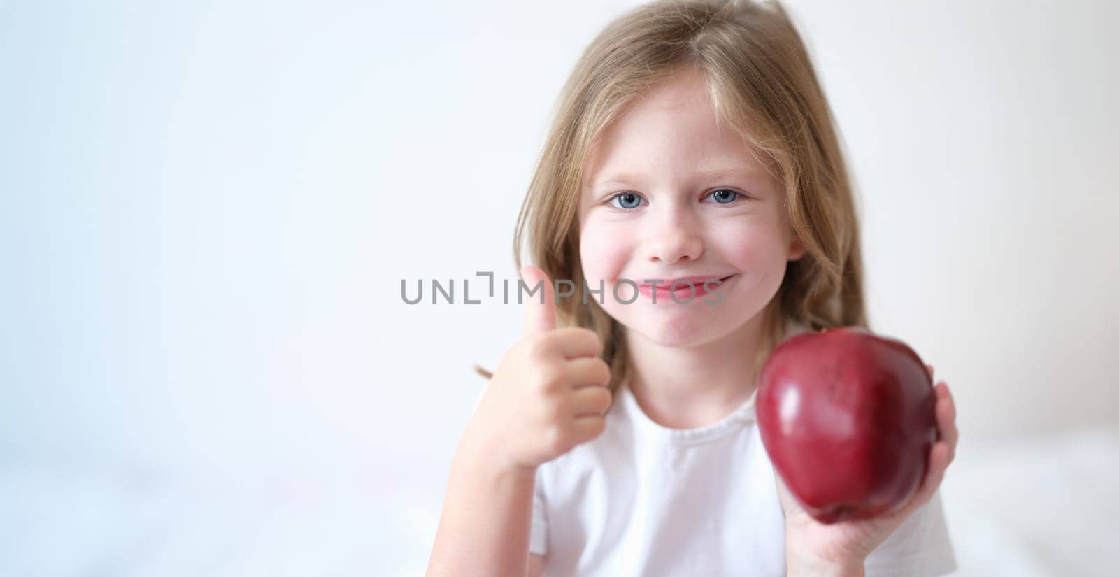 Portrait of little girl holding red apple in hands and showing thumbs up gesture. Healthy food and vitamins