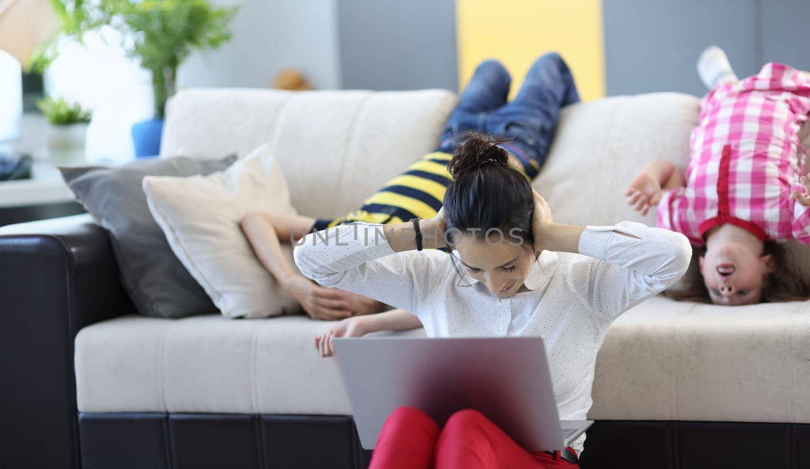 Portrait of tired mom trying to concentrate and work kids going crazy and jump. Woman closed ears with hands. Remote job, work from home and parenthood