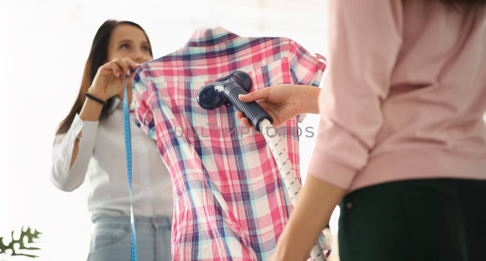 Close-up of atelier women using clothing steamer on plaid shirt, prepare product for selling. Females ironing blouse. Fashion and sewing concept
