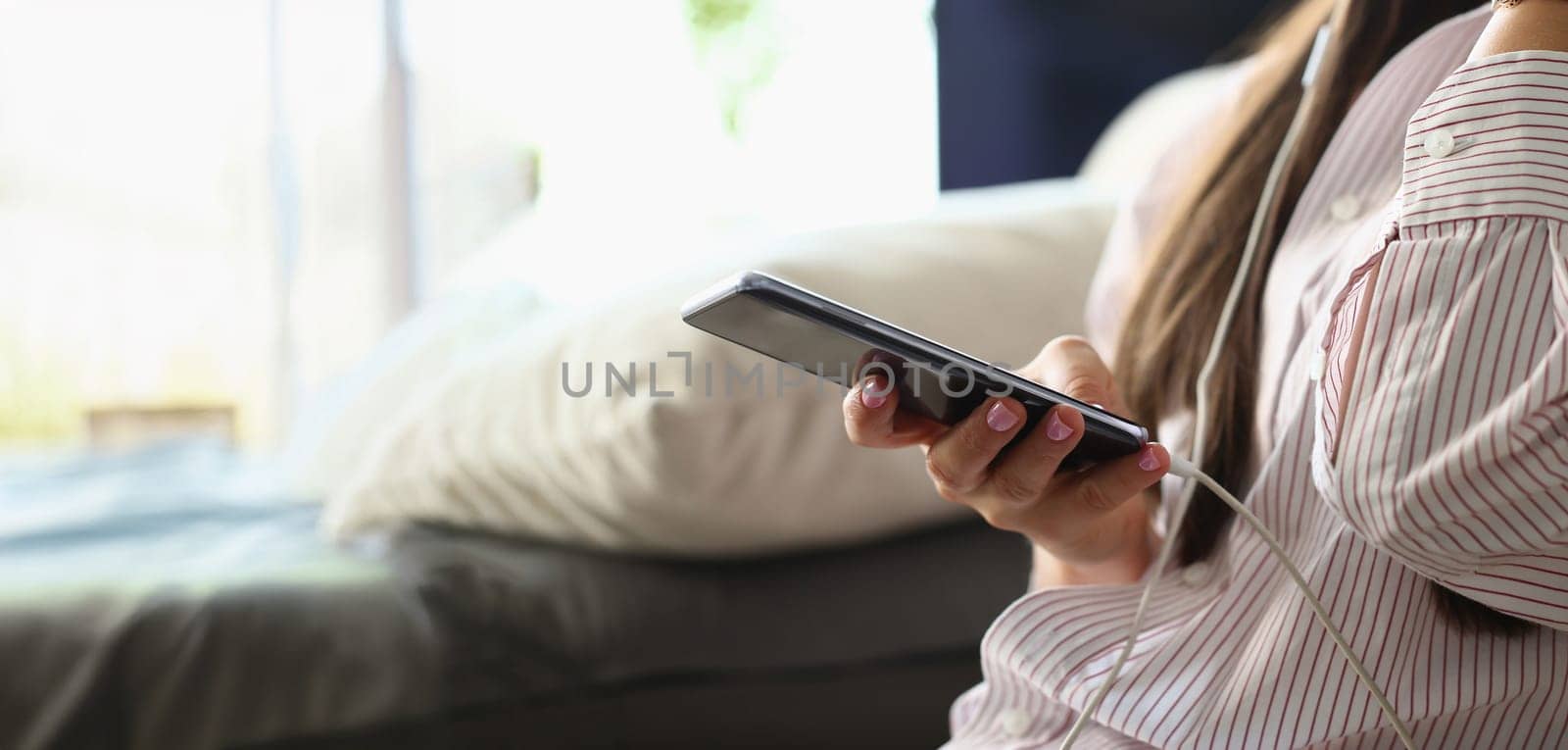 Close-up of female sitting on couch with smartphone and listening music. Woman watching film, surfing internet or messaging. Social media and entertainment concept