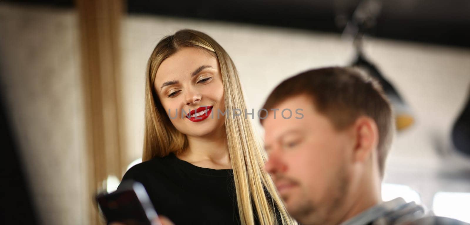 Portrait of smiling pretty blonde showing photo in smartphone to man. Cute stylish woman with fashionable makeup communicating with man. Red lips and beauty