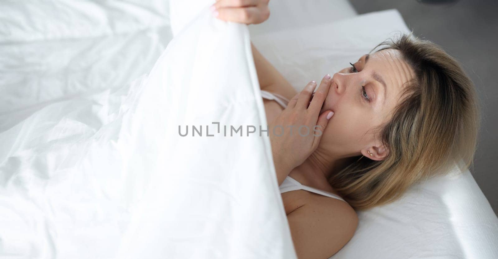 Top view of shocked surprised woman covering face with hand and looking under blanket. Heavy menstruation or unexpected surprise concept