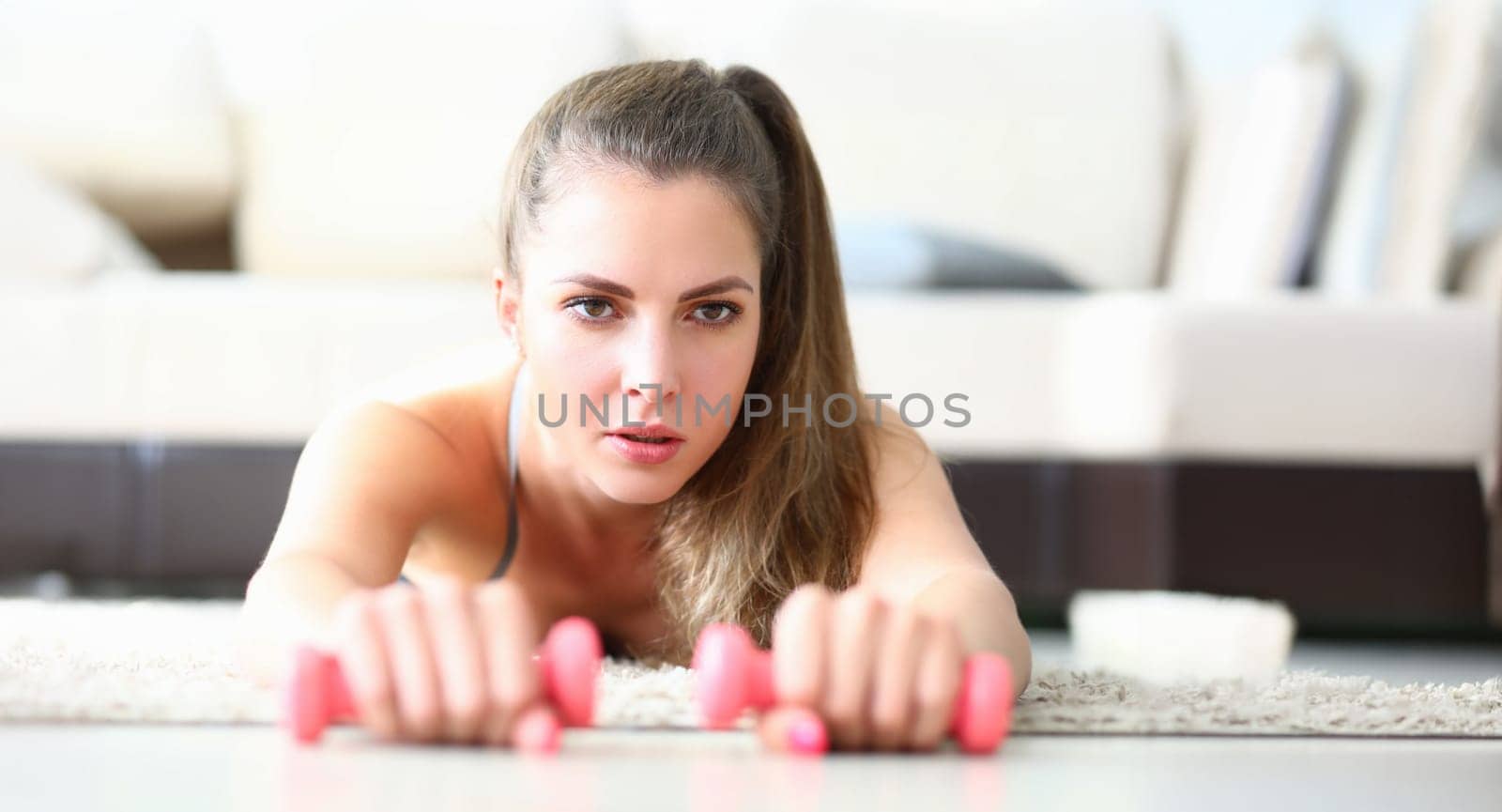 Portrait of cute young woman performing exercise with dumbbells. Fit female trainer training body with sport equipment at home. Sporty lifestyle and hobby