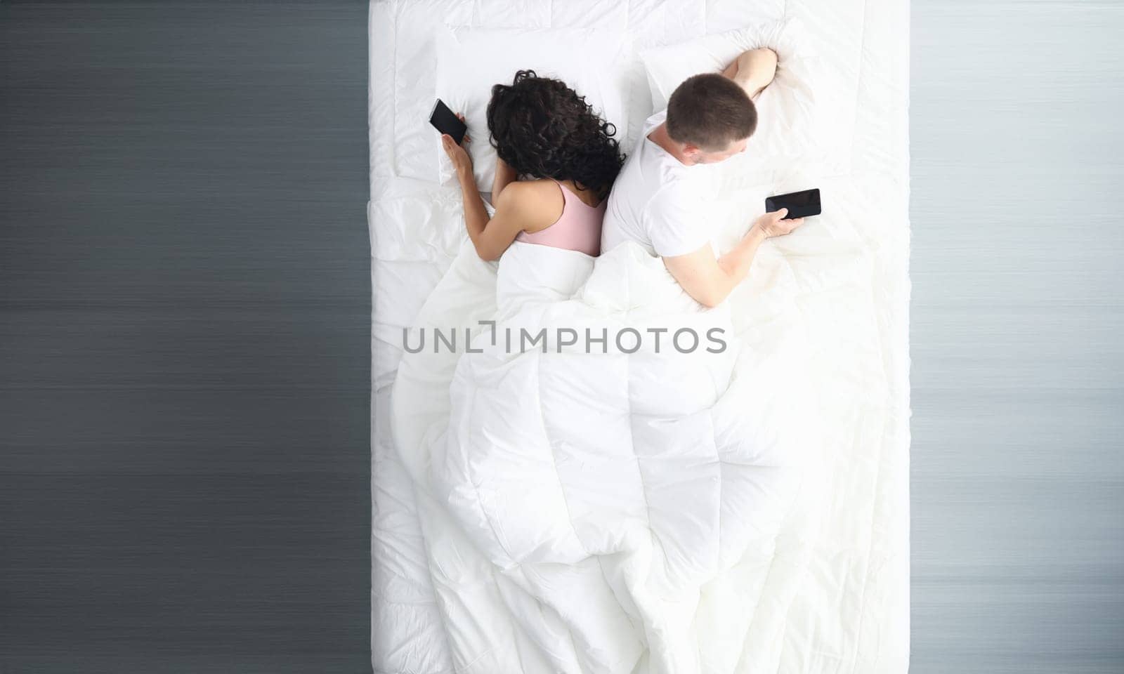 Top view of couple in bed using phones lying backs to each other. Man and woman surfing internet using cellphones. Relationship problems and virtual life