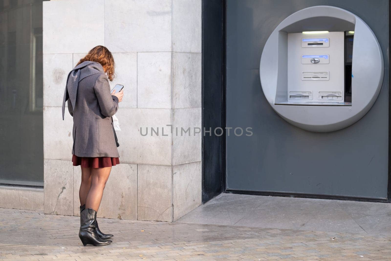Urban dweller in stylish attire engrossed in smartphone near an outdoor ATM. Woman at Bank using smartphone. download image