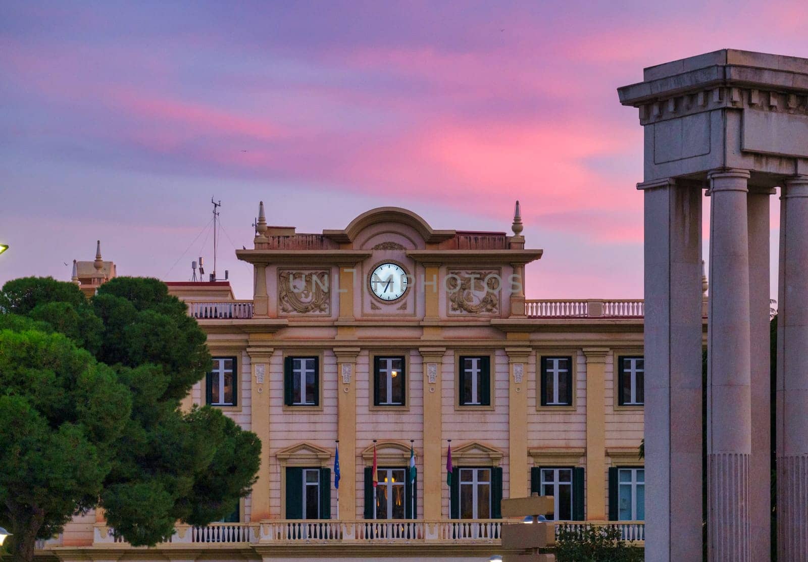European-style Spain building with clock, bathed in warm sunrise sunset light, amidst green trees and grand architecture. download image