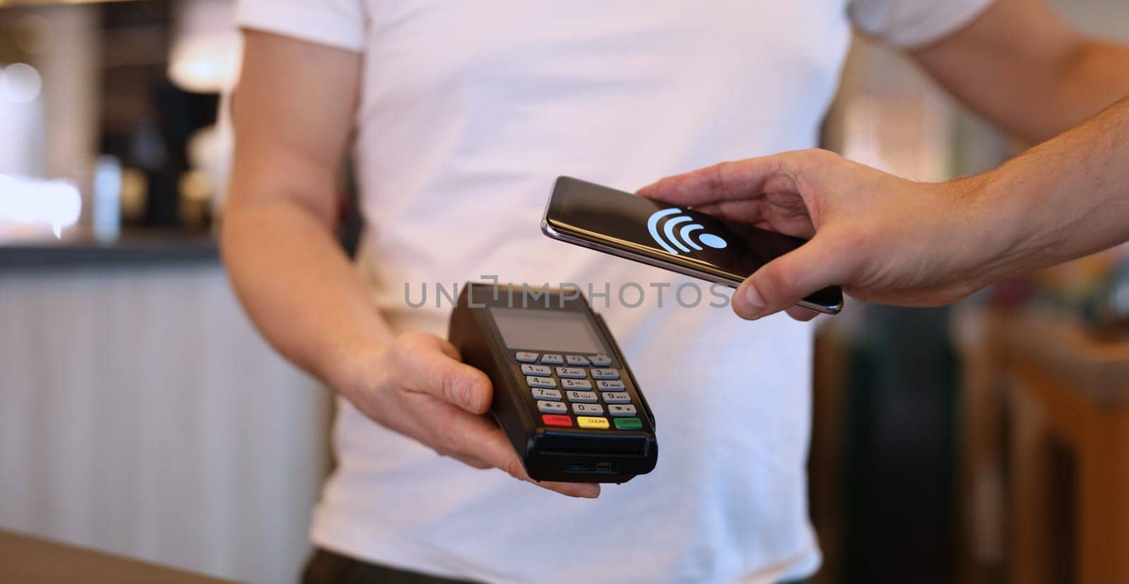 Close-up of man paying with mobile phone over pos terminal with nfc technology. Man using smartphone for wireless money transfer. Customer paying bill in cafe