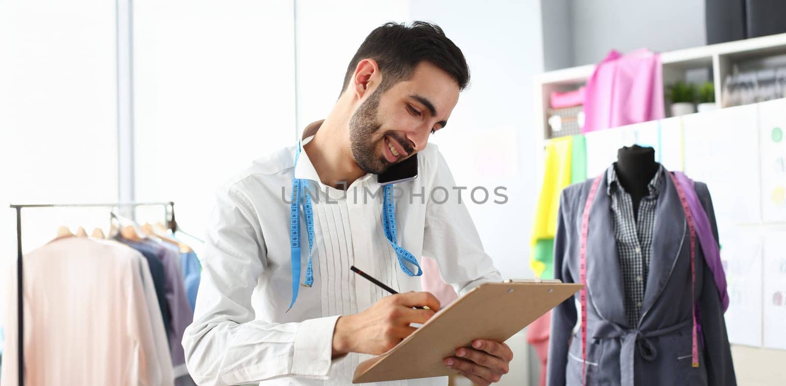 Smiling man tailor speaking on smartphone with client. Designer taking new order in atelier. Sewing workshop or creating new brand concept