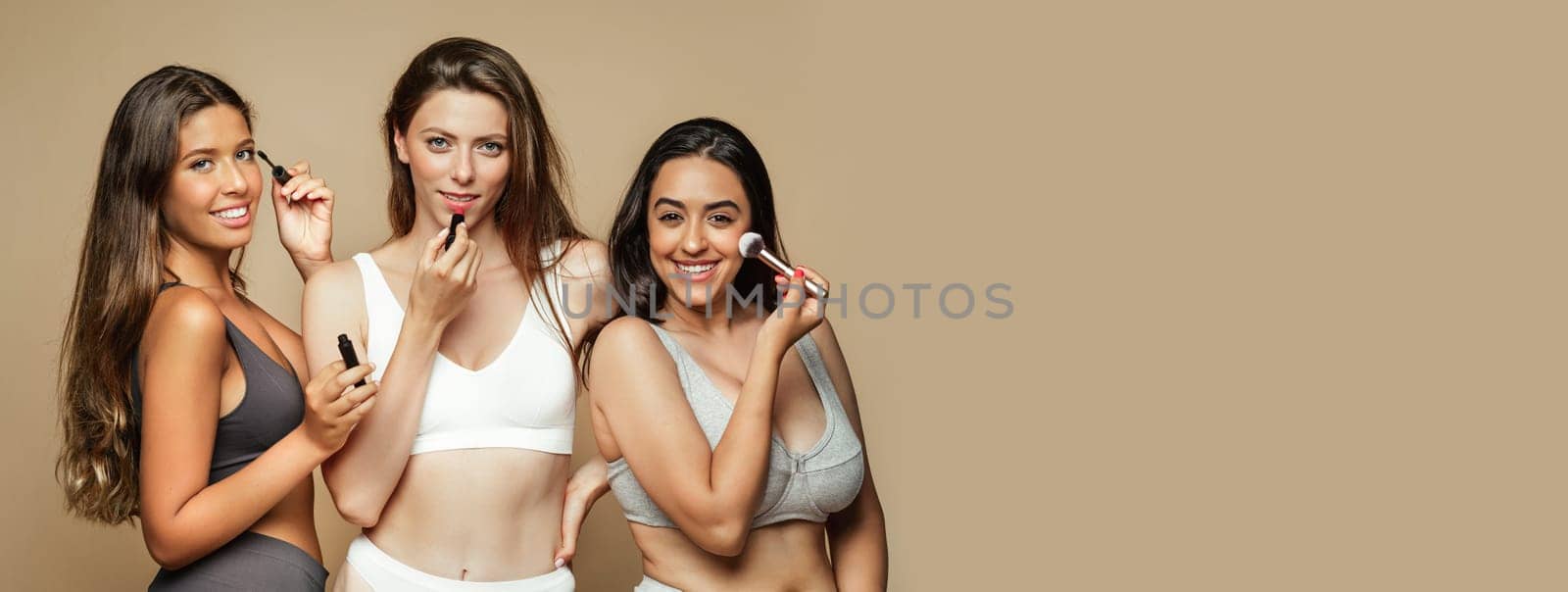 Happy young arabic, spanish and european women in different underwear enjoy natural beauty with brushes and lipstick, isolated on beige background, studio. Cosmetics, nude makeup, routine, copy space