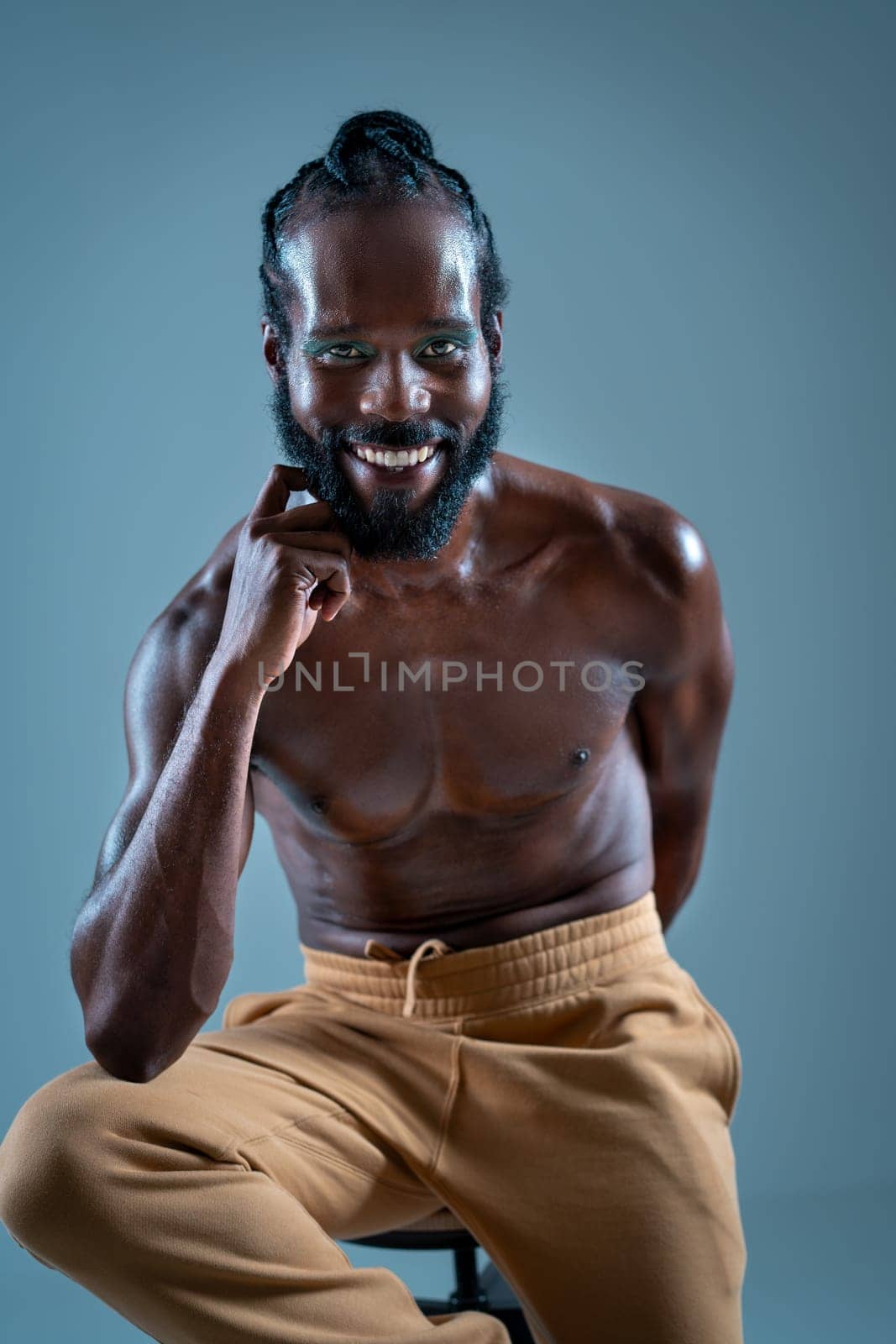 Smiling African American gay man with bright makeup. Shirtless muscular man sitting on stool and looking at camera. LGBTQ person is posing on blue background.