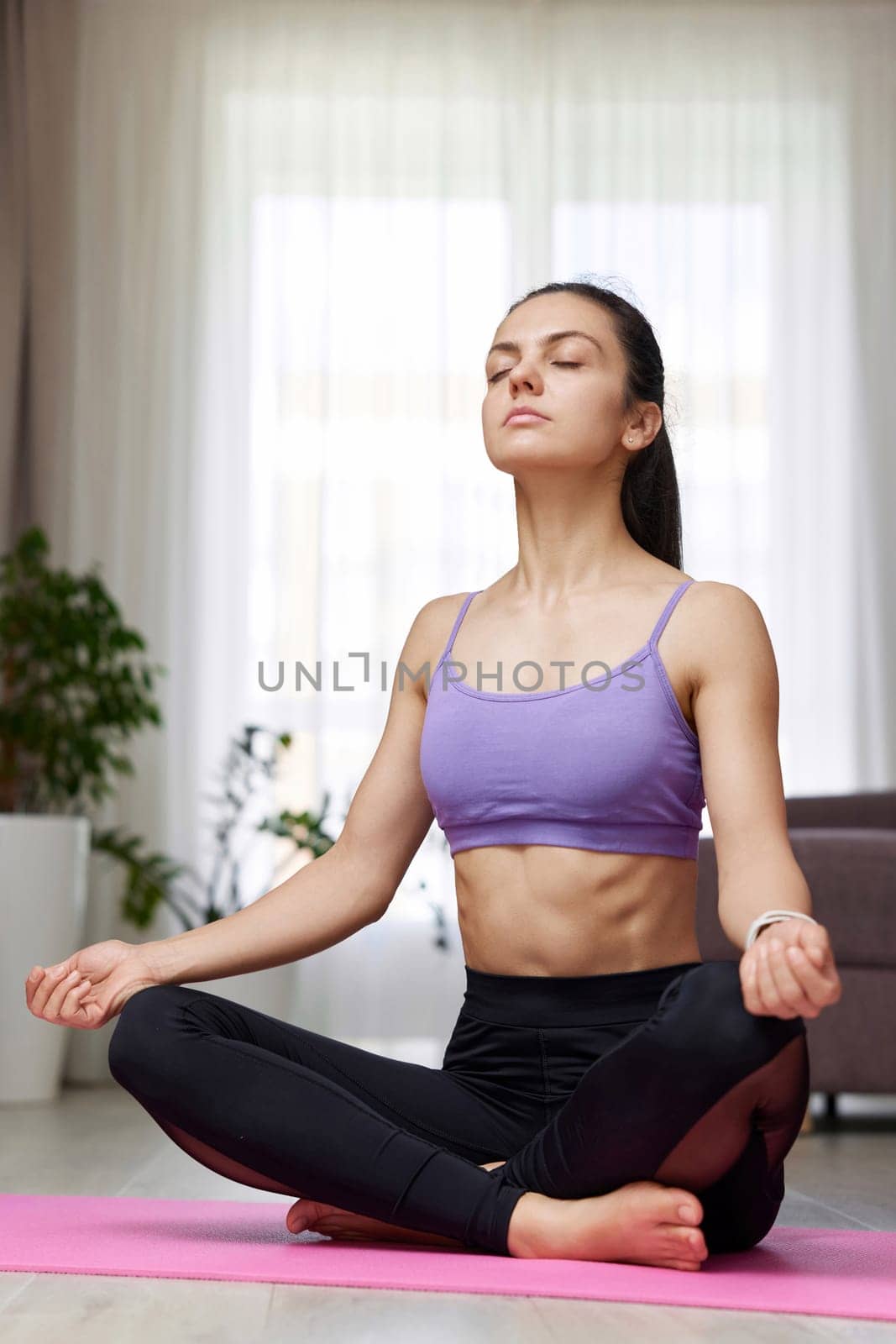 Caucasian woman with closed eyes practicing yoga and meditation sitting in lotus pose on pink yoga mat at home