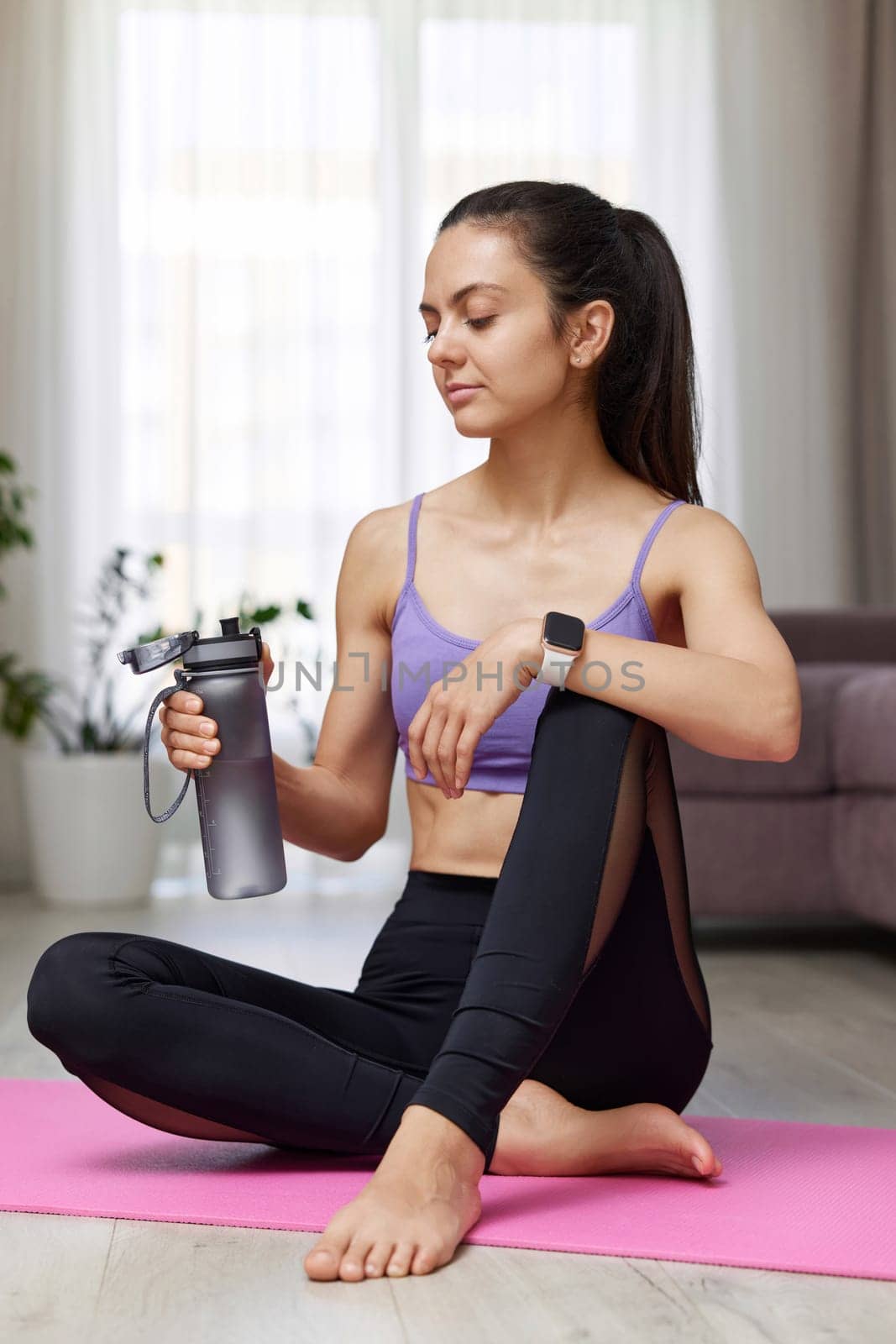 athletic fitness woman drinking water sitting on pink exercise mat at home