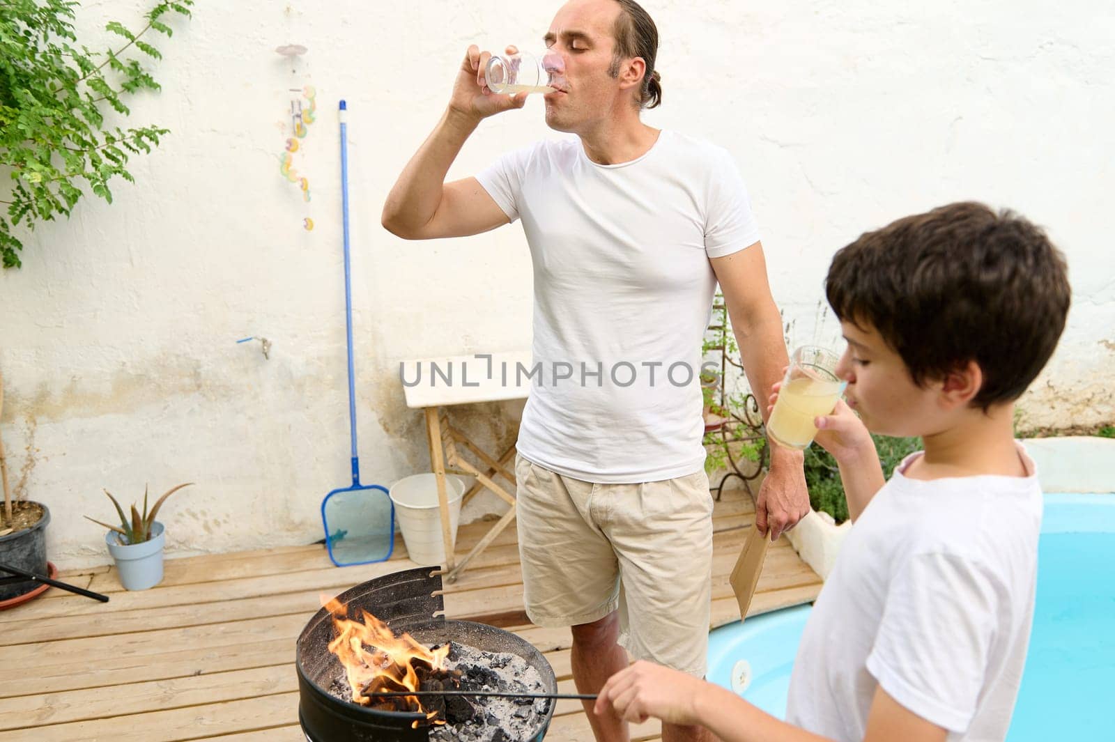 Father and son bonding during a summer barbecue by the pool. The father is drinking water while the son sips lemonade. Perfect family time and outdoor cooking concept.