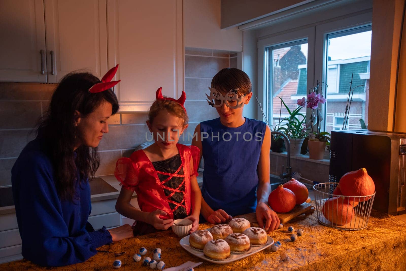 Mother, daughter and son in Halloween costumes draw skulls with cocoa powder and powdered sugar on sweet buns in a darkened room, baking together a traditional dessert for the holiday