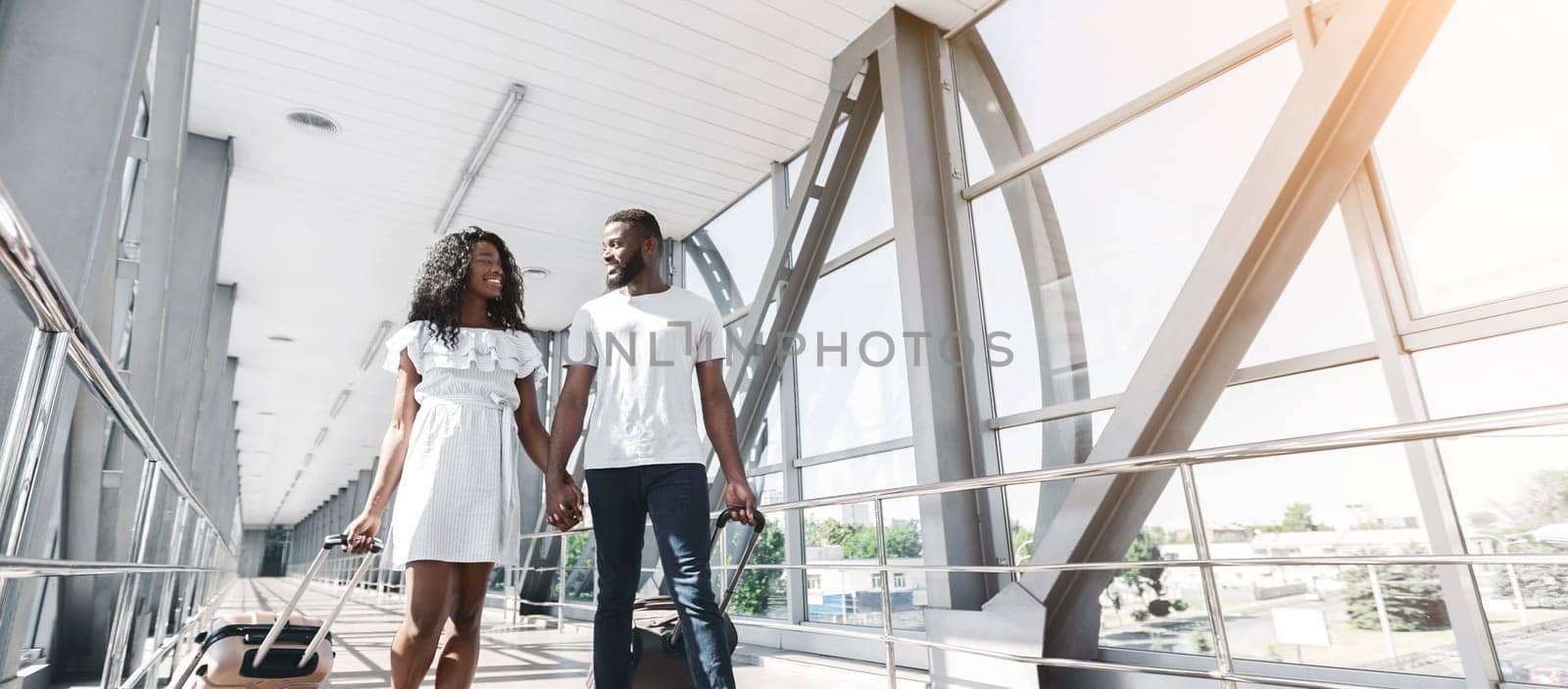 African American young couple walks hand-in-hand through an airport terminal, each pulling a wheeled suitcase. They are smiling and looking at each other, excited for their upcoming trip