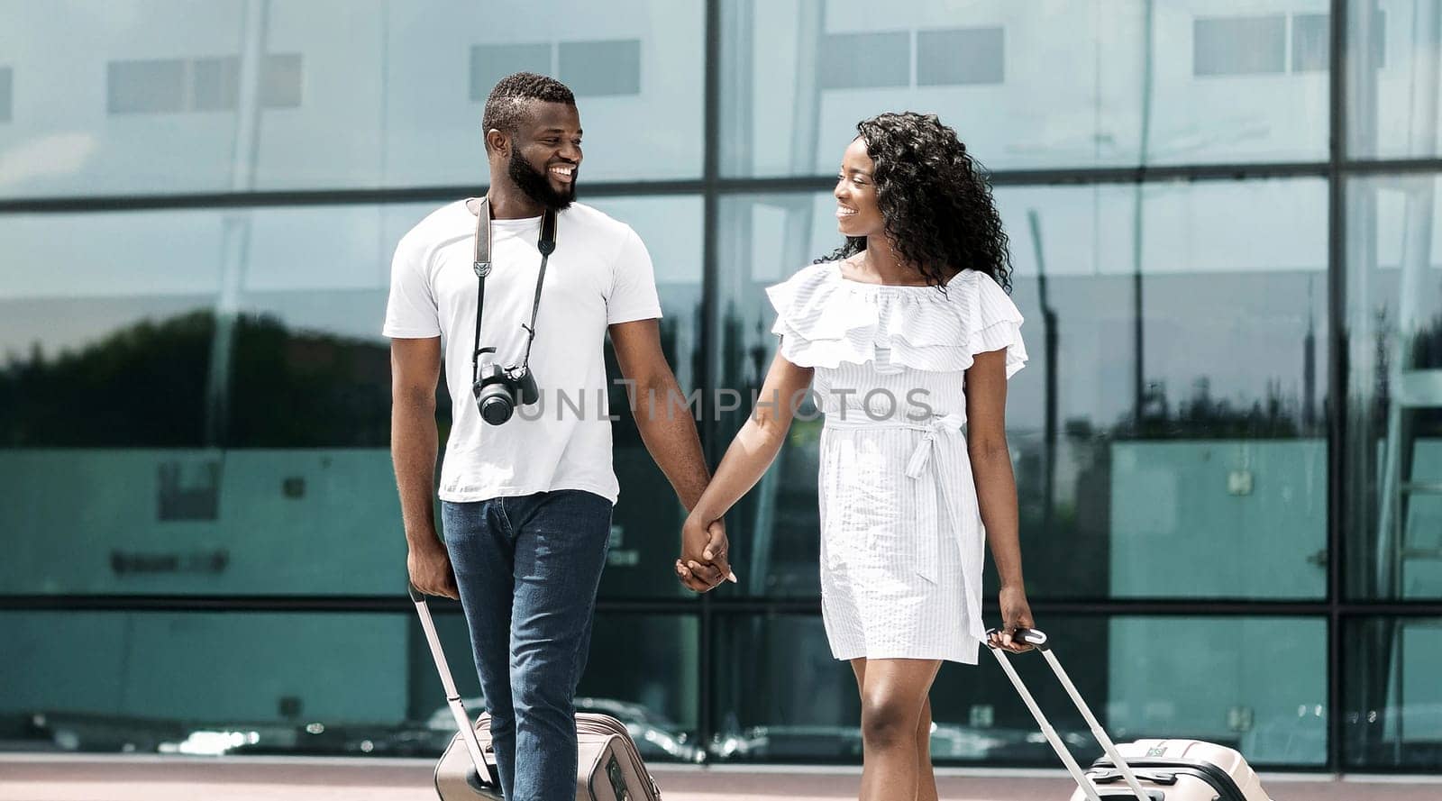 A happy black couple walks through an airport terminal, holding hands and pulling their luggage behind them. Traveling, tourism