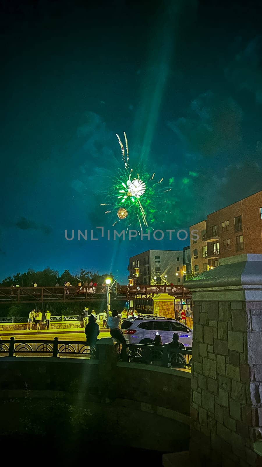 Castle Rock, Colorado, USA-July 4, 2024-A vibrant July 4th fireworks display lights up the night sky, with bright green and gold bursts creating a stunning visual against the dark backdrop. The scene captures the excitement and celebration, with spectators gathered in a suburban area featuring modern buildings and festive lights. The festive atmosphere is palpable as people enjoy the patriotic celebration.