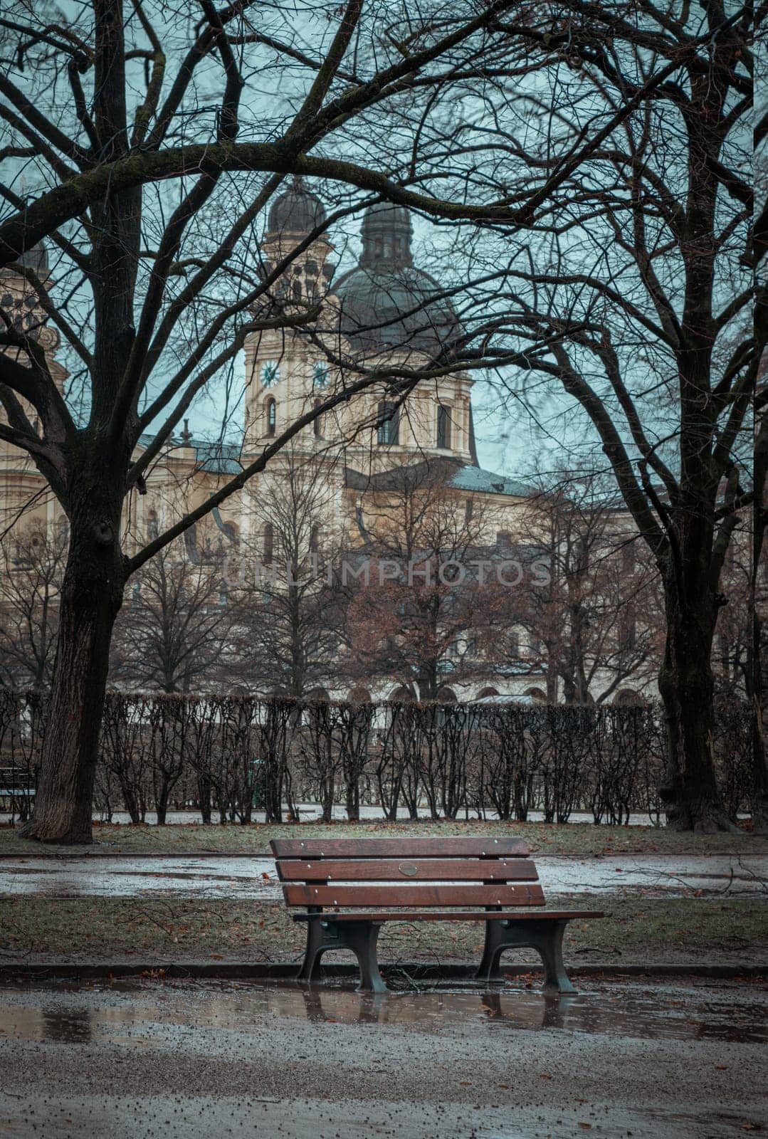 Munich, Germany - Dec 22, 2023 - Bench made wood and iron stand in the garden of the Munich Residenz with St.Cajetan and Adelaide in the background. Space for text, Selective focus.