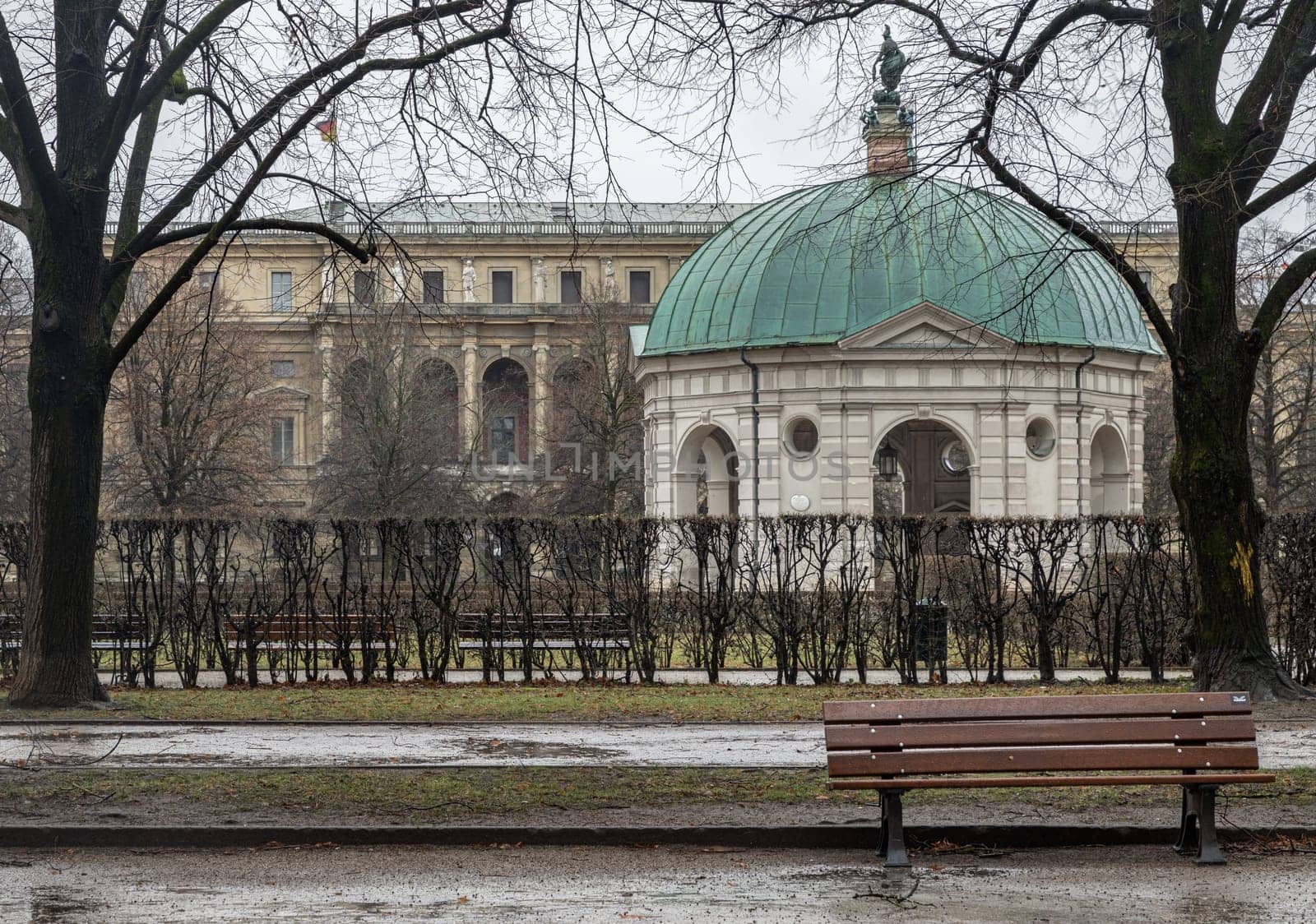Munich, Germany - Dec 22, 2023 - Bench made wood and iron stand in the garden of the Munich Residenz with The Diana Temple (Dianatempel) and The Munich Residence Museum Building in the background. Space for text, Selective focus.
