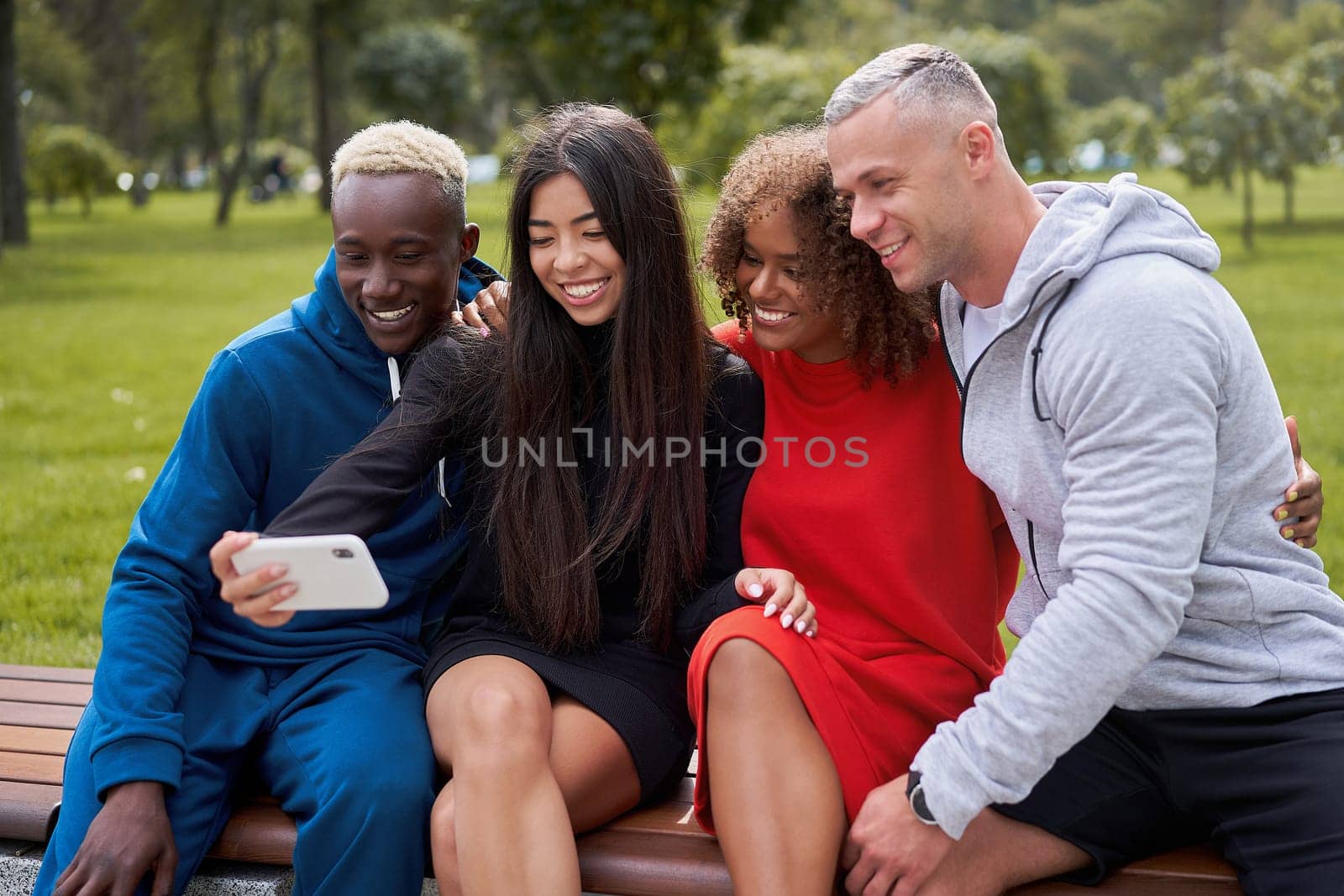 Smiling multi-ethnic friends taking selfie on smartphone. Diverse group people spending time together sitting on bench. Multiracial male female students in colorful sportswear during weekend.