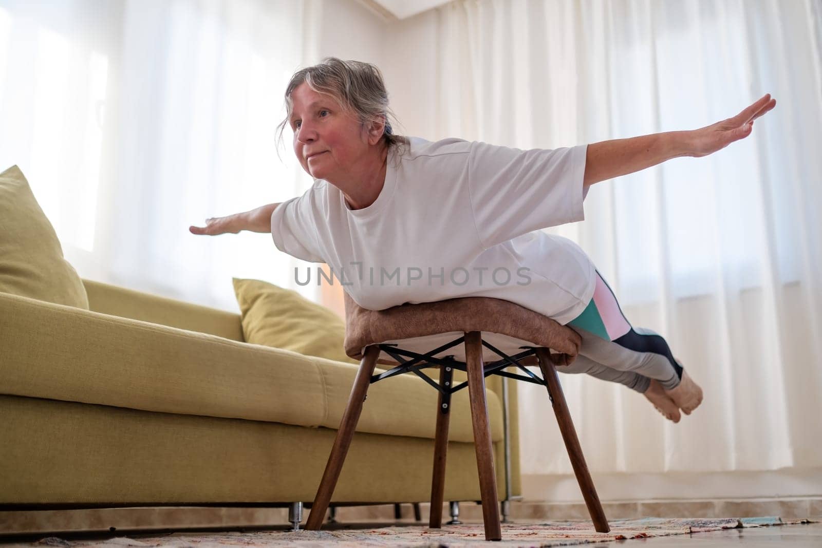 Senior caucasian woman doing yoga at home using chair as a props.