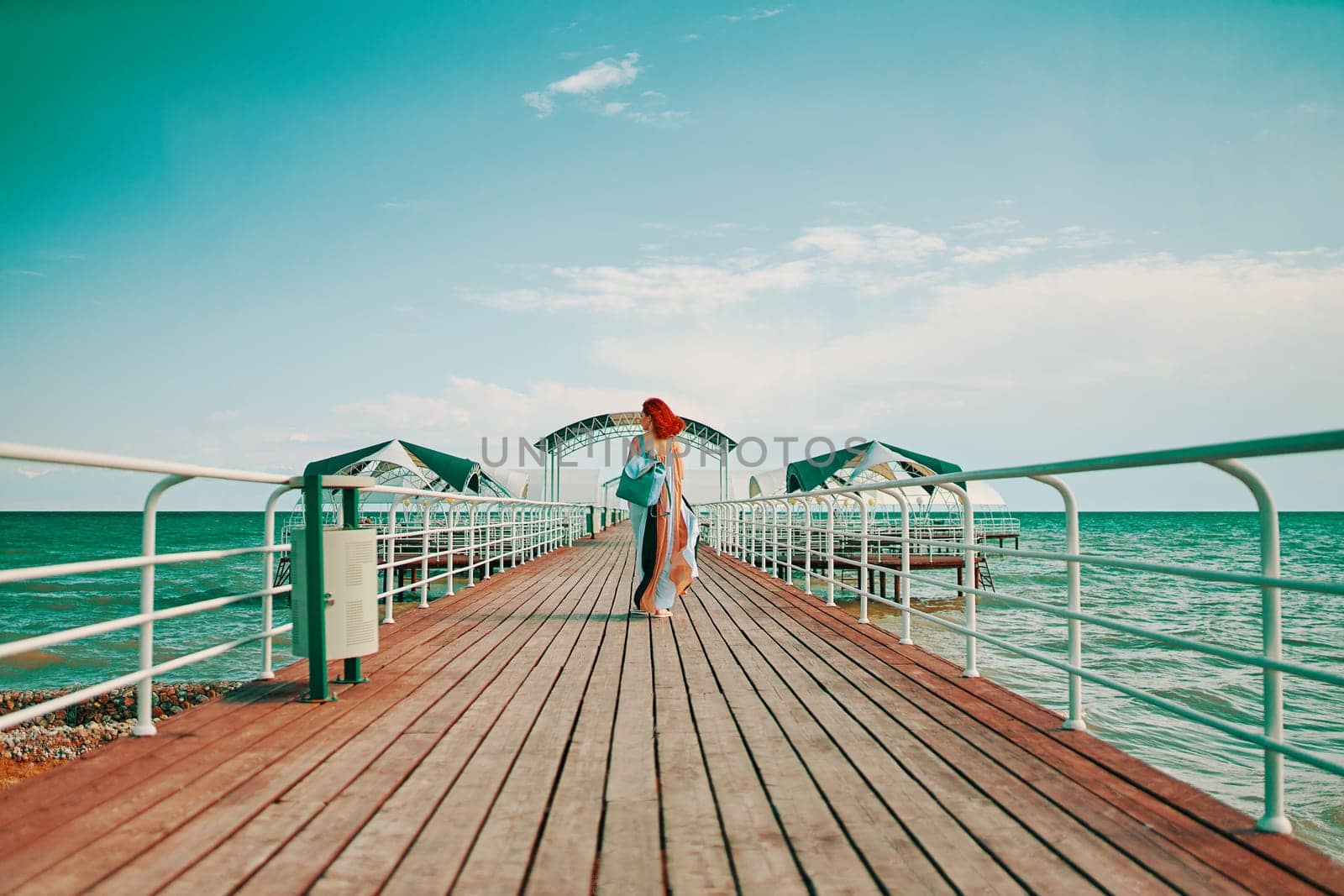 The resort pier is graced by the presence of a red-haired beauty in a summer dress, accompanied by a backpack, as she enjoys a serene walk with the sea in view.