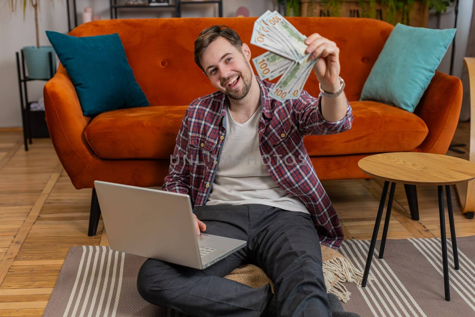 Planning budget. Rich happy excited Caucasian man counting money cash dollar using laptop calculate domestic bills at home. Young guy satisfied of income earnings, saves money for planned vacation.