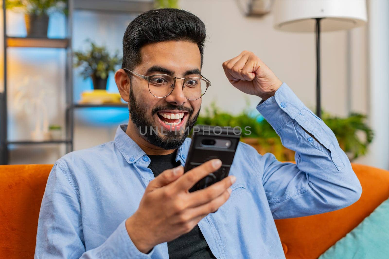 Surprised excited Hispanic young man winner holding smartphone reading good news amazed by online bet bid game win. Happy Indian guy on sofa looking at screen overjoyed by victory success at home.