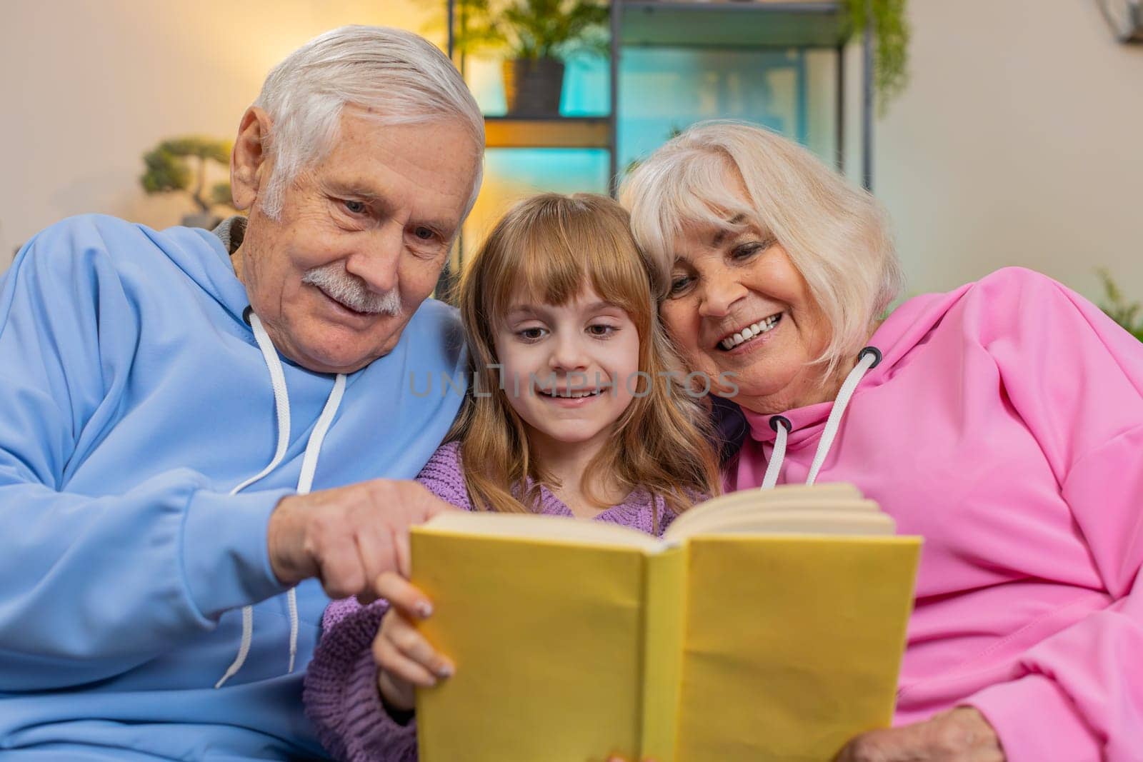 Caucasian grandparents and cute granddaughter reading interesting book together sitting on sofa in living room at home. Girl with grandparents spending leisure time on comfortable couch in apartment.