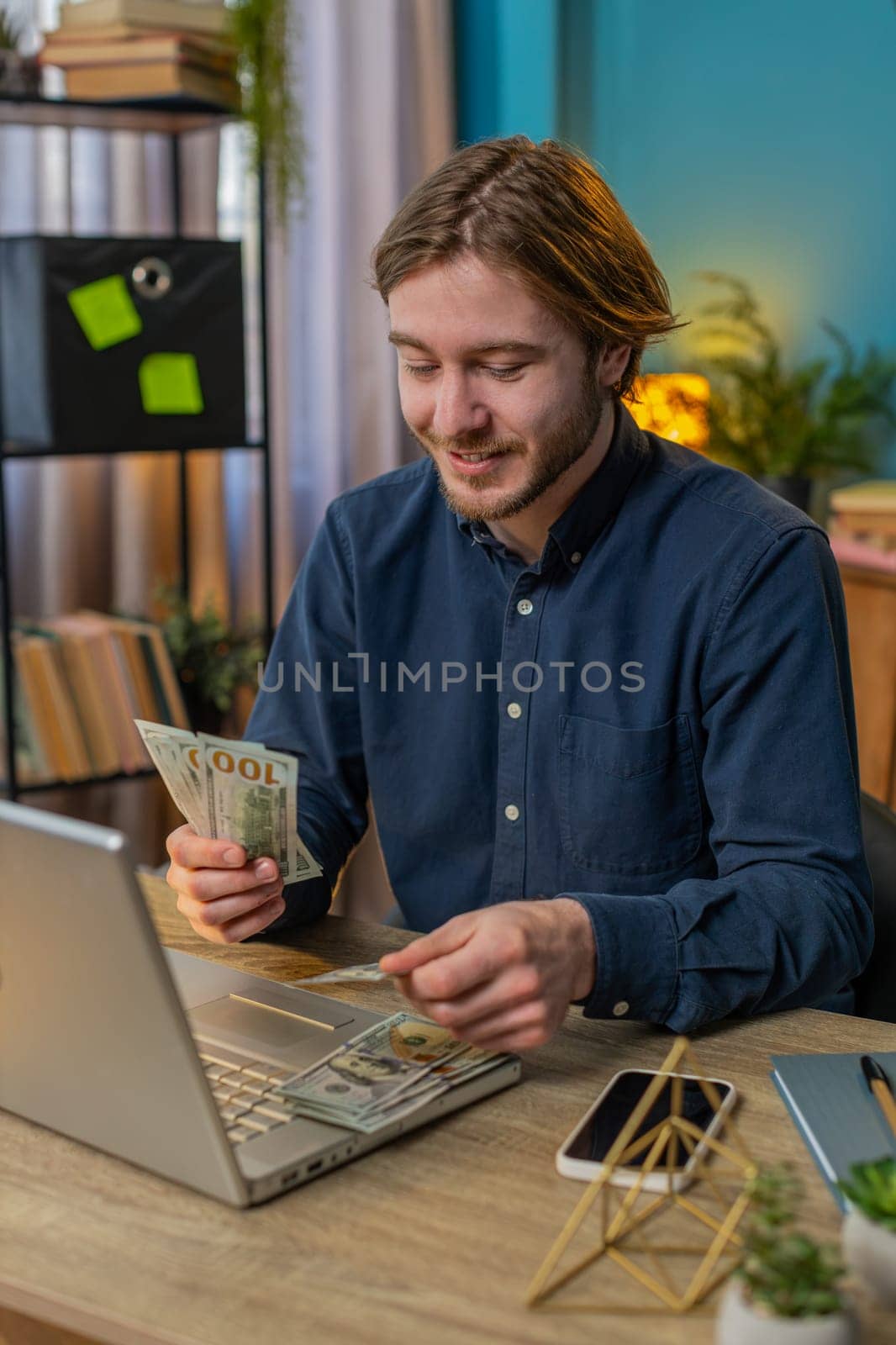Businessman holding fan of cash money dollar banknotes showing thumbs up and pointing at laptop, celebrate, success career, online income, wealth at home office desk. Remote distant working. Vertical