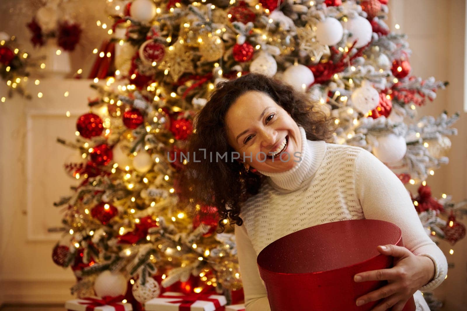 A happy woman holding a red gift box during the Christmas season, with a beautifully decorated tree and festive ornaments in the background. Celebrating the holiday spirit and joy.