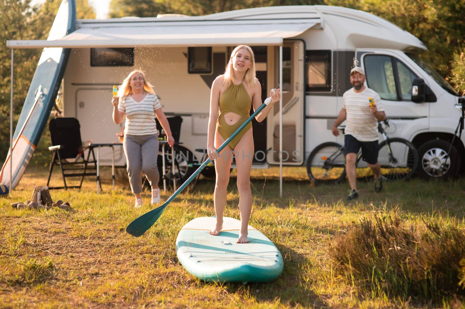 the family is resting next to their mobile home. My daughter is standing with a paddle on a sup board, and her parents pour water on her.