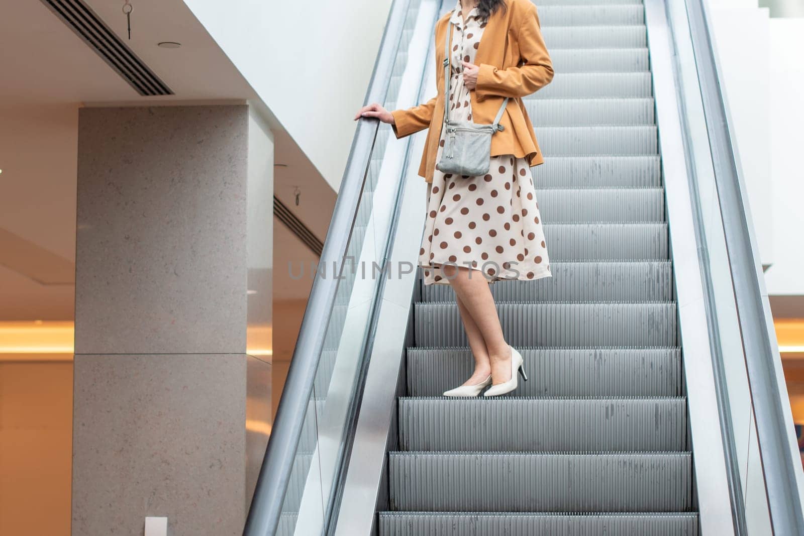 Unrecognizable young woman in a beige dress with large polka dots goes down an escalator in a shopping center, blurred background