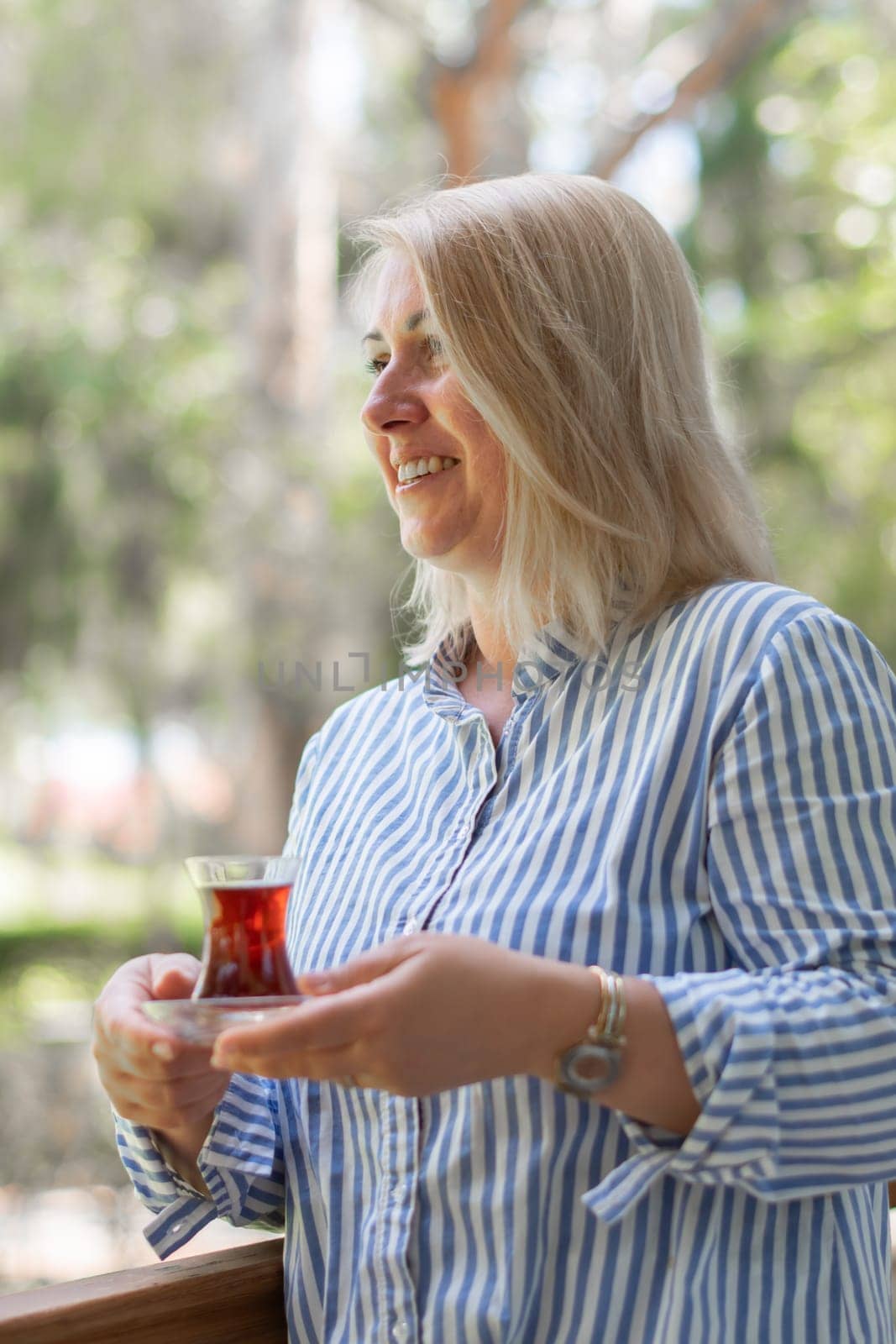 young blonde Caucasian woman drinks tea from Turkish Armudu cup on the balcony and takes bite of orange cake, delicious dessert after lunch, traditional oriental sweet on summer vacation