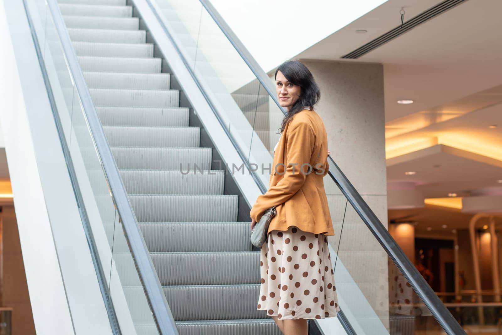 Confident young brunette goes up the escalator to the floor above in a shopping center, looks at the camera, blurred background, High quality photo