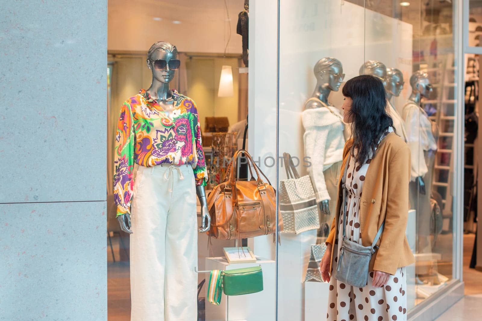 Confident young brunette looks dreamily at a display case with luxury bags and mannequins in elegant clothes in a boutique of modern shopping center, High quality photo