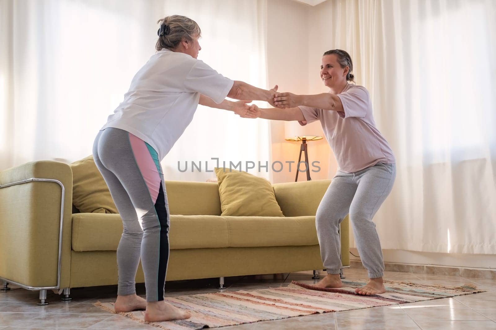 Senior woman exercising at home together with daughter. Active mature woman doing stretching exercise in living room at home.