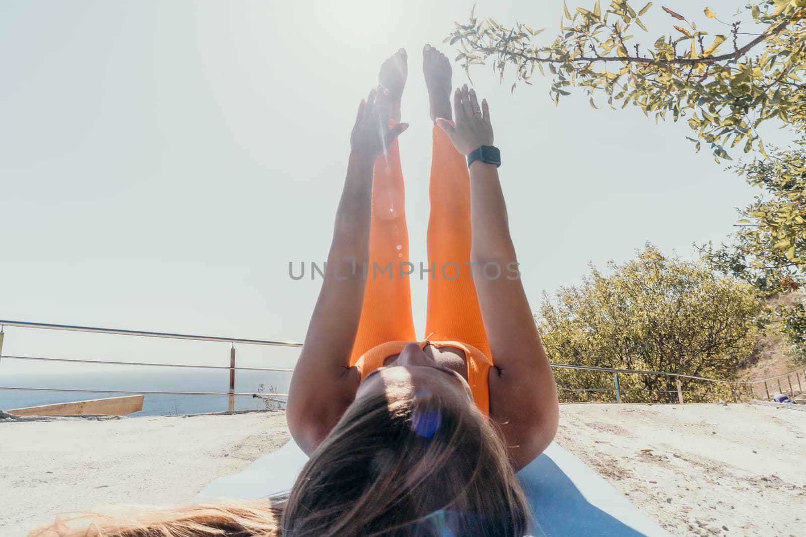 A young woman in orange activewear performs a leg raise exercise on a yoga mat outdoors. She is enjoying the view while working out.