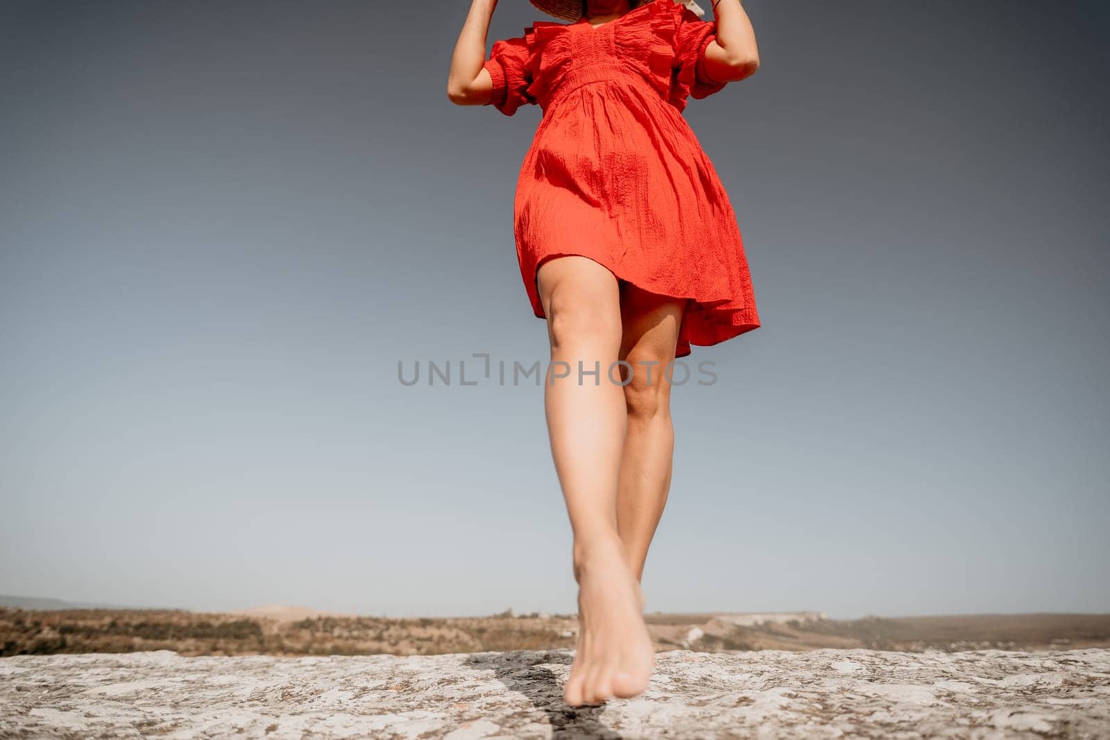 A woman in a red dress walks barefoot towards the camera, a cliff and blue sky in the background.