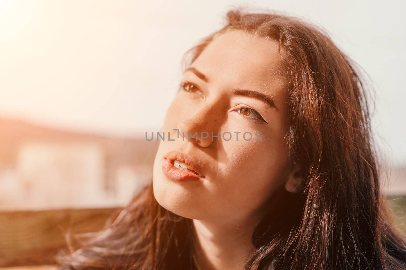 Close-up portrait of a young woman with brown hair looking upwards, captured on a sunny day, highlighting the golden light on her face.